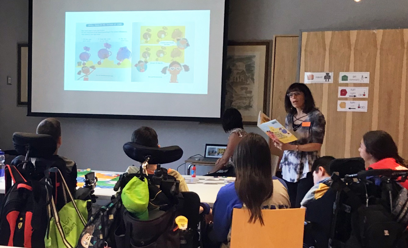 Reading picture books before the start of an activity to help set the context for the collaborative tasks Woman reading a book for a group of children on mobility devices and projecting the page on the screen.